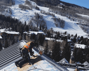 A construction worker in a safety vest kneels on a steep roof, using a tool, with snowy mountains, trees, and buildings visible in the background under a clear blue sky.