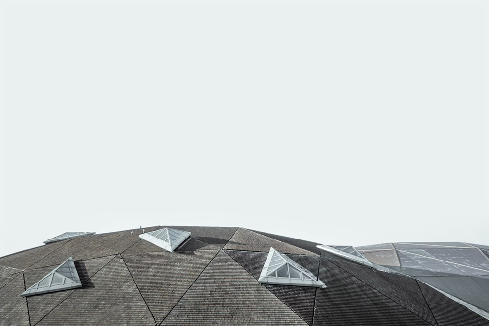 A minimalist view of a large, modern, geometric roof with angular, triangular facets integrated with window segments. The sky is overcast, providing a muted backdrop that accentuates the architectural design.