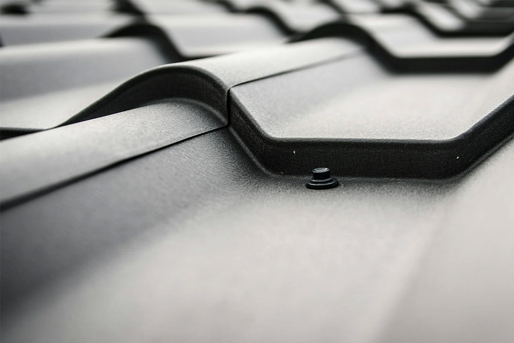 Close-up view of a gray, metal tiled roof showcasing the texture and contours of the roofing material. The image highlights the overlapping design and a visible screw, indicating the fastening method used to secure the metal tiles.