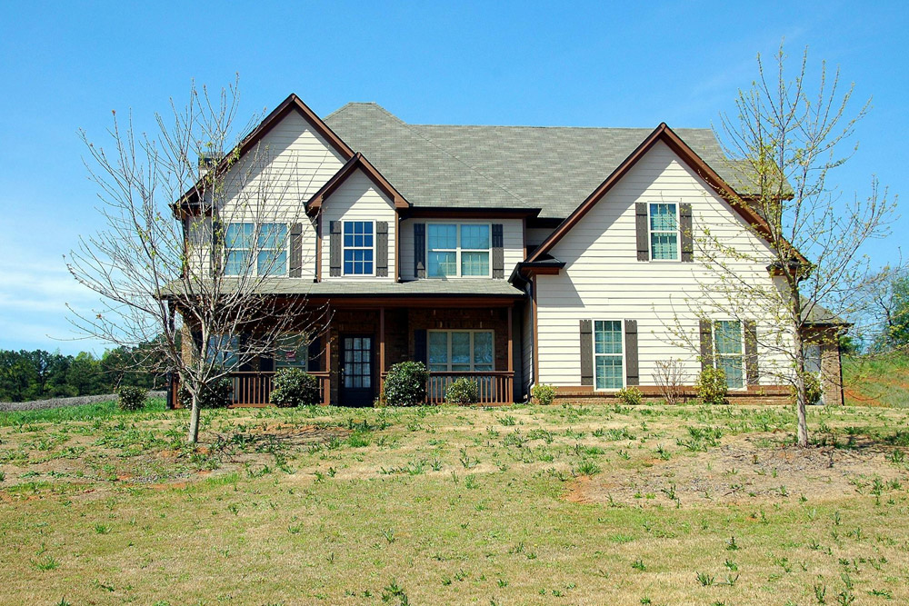 A large two-story house with beige siding and dark trim sits on a grassy lot. The front porch is surrounded by small shrubs and young trees. The sky is clear and blue, enhancing the serene countryside setting.