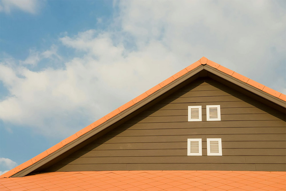 The image shows the top of a house with a triangular gable and a red-orange roof. The facade is brown with two sets of white shutters. The sky is partly cloudy with soft, white clouds.