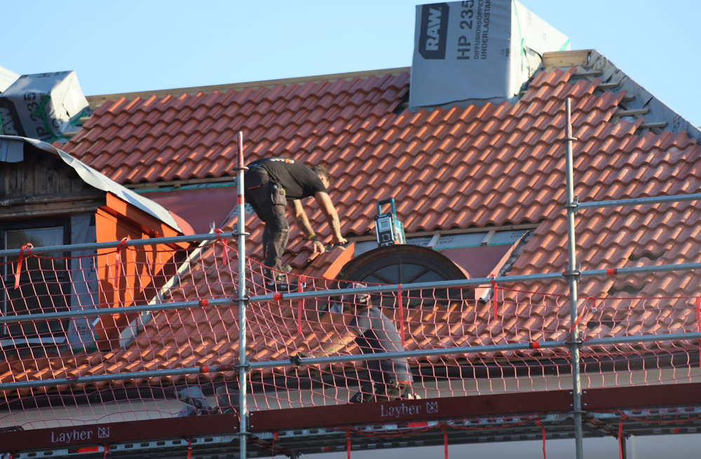 A worker stands on scaffolding and tiles a sloped roof with red shingles around a window on a building under construction.
