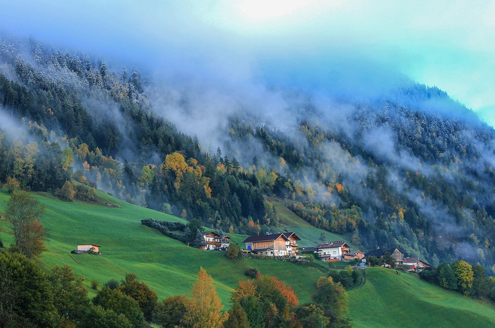 A small village with rustic houses sits on green hills, surrounded by colorful autumn trees, mist, and forested mountains partially covered in fog.