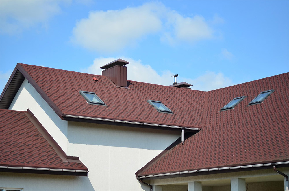 A modern house roof with brown shingles, several skylights, and a small chimney against a blue sky with scattered clouds.